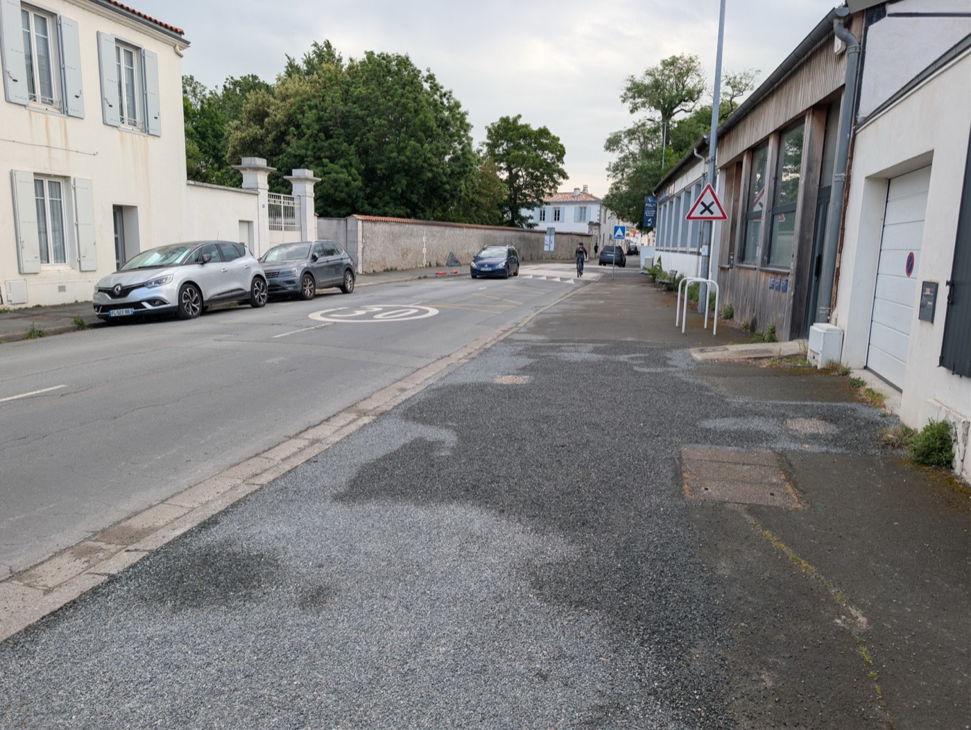 Rue Marius-Lacroix le 30 avril 2026, vue d'ensemble : marquage zone 30 au sol, gravillons et asphalte rapiécé sur la chaussée, voitures stationnées à gauche, cycliste