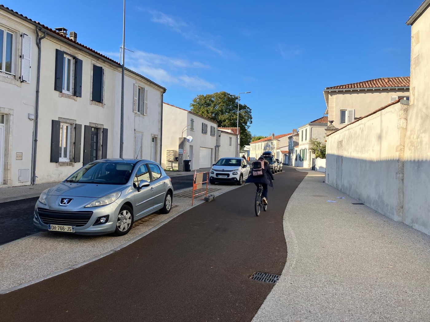Une cycliste emprunte la piste cyclable achevée entre le trottoir refait et des voitures stationnées sur une bande stabilisée